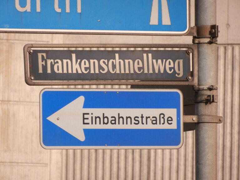 Foto eines Schildermastes drei links montierten Verkehrsschildern, dahinterliegender Betonwand. Oberstes Schild Hinweis zu einer Autobahn, in der Mitte das Straßenschild "Frankenschnellweg", darunter das Verkehrszeichen "Einbahnstraße (nach links)"