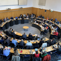 Besprechungssaal mit kreisförmig angeordneten Tischen, an denen Stadträtinnen und Stadträte sitzen.
