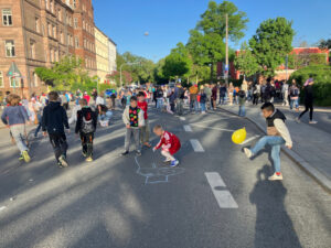 Kinder, die vor der Schule auf der autofreien Straße sicher spielen. Im Vordergrund ein Junge, der einen gelben Luftballon kickt.