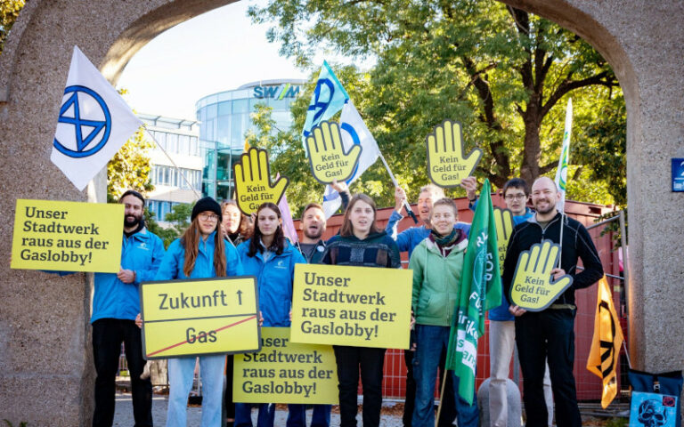 Eine Gruppe Menschen mit Plakaten "Unsere Stadtwerke raus aus der Gaslobby!" unter einem Tordurchgang
