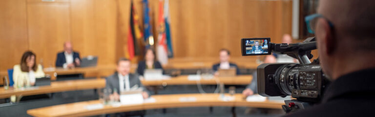 Teilnehmende des Bürgerdialogs im Sitzungssaal des Stadtrats - unscharf. Vorne rechts (scharf zu sehen) ein Kameramann.