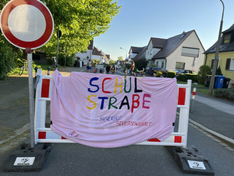 Eine Absperrbaake mit einem übergeworfenem Laken, auf dem in bunten Buchstaben steht "Schulstraße - für Sicherheit im Straßenverkehr und Selbständigkeit. Links davon Verkehrzeichen 250 - "Verbot für Fahrzeuge aller Art"