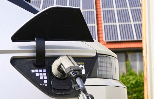 Bild mit der Ladebuchse eines E-Autos mit eingestecktem Ladekabel. Im Hintergrund Photovoltaik-Panelle auf einem Hausdach.