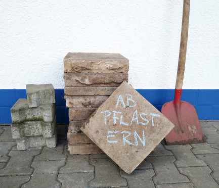 Vor deiner Wand gestapelte Pflastersteine und eine Schaufel. Ein Pflasterstein steht auf der Ecke - darauf steht mit Kreide "Abpflastern".